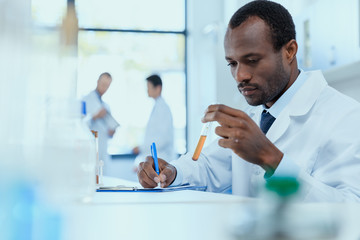 African american scientist in white coat holding and examining test tube with reagent, laboratory...