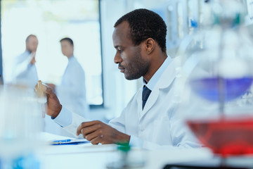 Side view of african american scientist in white coat holding and examining test tube with reagent,...