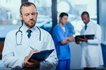 portrait of confident doctor writing in folder in clinic with colleagues behind