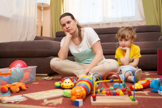 Young Pregnant Mother And Her Little Son, At Home. Mother And Little Son  Sit On A Floor. Mom Tired To Tidy Up The House. Child Scattered Toys. Mess In The House. Mother Has Quarreled With The Son.