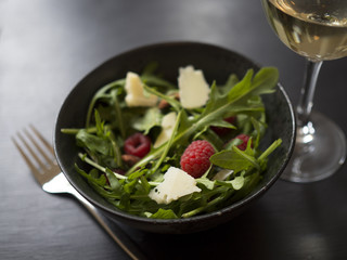 Fresh rocket salad with raspberries, roasted almonds and cheese in black ceramic bowl, and a glass of white wine on black wooden table