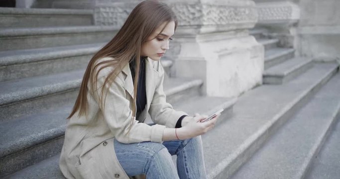 Side view of young female wearing coat and jeans and sitting on stairs of old building using smartphone