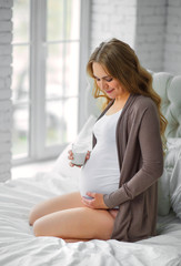 Portrait of a beautiful pregnant woman sitting on bed with glass of milk. Breakfast. The concept of useful diet pregnant. Side view.