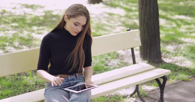 Beautiful young woman in casual clothing sitting on bench alone and using tablet on blurred background of park