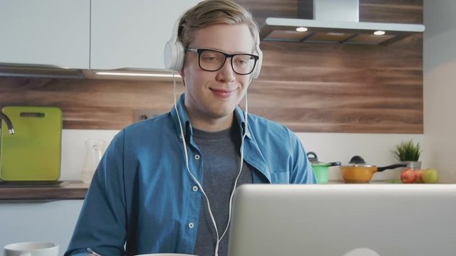 Young laughing man watching streaming movie on laptop at home eating cereals with headphones