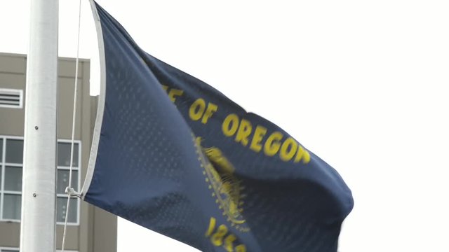 A State of Oregon flag waving in strong winds on an overcast day. corner  of a building is viable in the background.