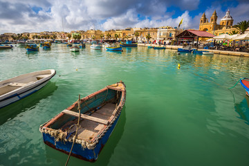 Marsaxlokk, Malta - Traditional colorful maltese Luzzu fisherboats at the old market of Marsaxlokk with green sea water, blue sky and palm trees on a summer day