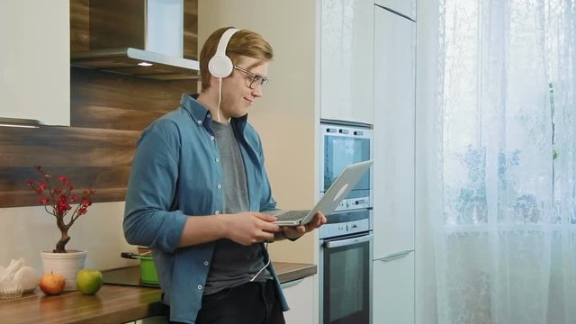 Young laughing man watching streaming movie on laptop at home eating cereals with headphones