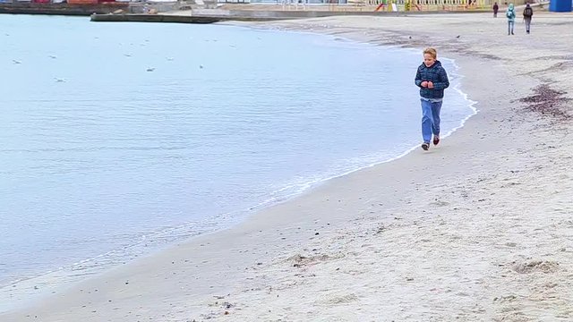 Little funny child runs fast to camera on autumn sea beach. Boy dressed in casual warm blue jacket and trousers. Seagull flying away. Real time hd video footage.