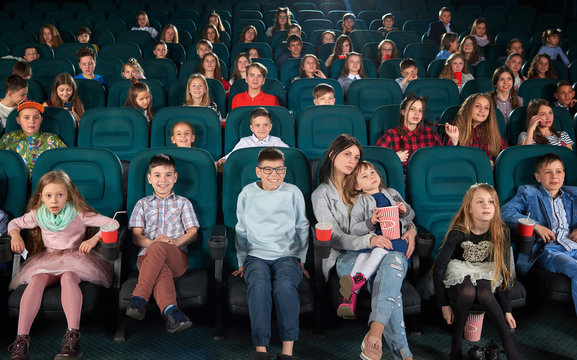 Full length shot of a woman enjoying watching a movie at the cinema with her little daughter in an auditorium full off children youth lifestyle leisure entertainment people activity concept. © serhiibobyk