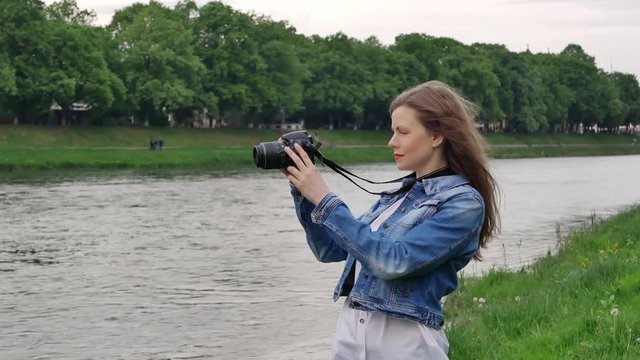 Beautiful girl tourist shoots a video with a professional camera on the banks of the river in windy weather