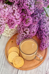 glass coffee Cup with milk on a wooden stand with cookies on a background of purple lilac bouquet