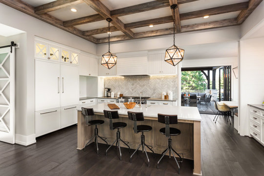 Beautiful Kitchen in New Luxury Home with Island, Cross Hatch Wood Beams on Ceiling, Hardwood Floors, Farmhouse Sink, and Elegant Pendant Lights. Open Doors in Background Lead to Outdoor Patio