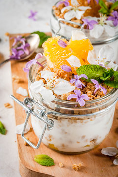 Trendy exotic food. Vegan paleo diet. Breakfast. Yoghurt with granola, fillet slices of orange, mint and edible flowers - lilac, cherry. On a white stone table, with ingredients. Copy space
