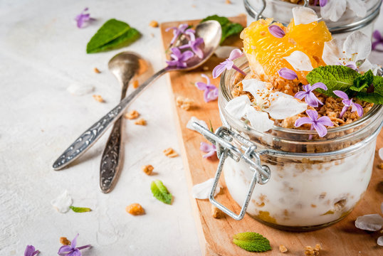 Trendy exotic food. Vegan paleo diet. Breakfast. Yoghurt with granola, fillet slices of orange, mint and edible flowers - lilac, cherry. On a white stone table, with ingredients. Copy space