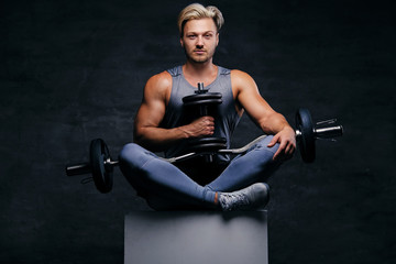 Fototapeta na wymiar The handsome blond sporty male sits on a white box with crossed legs and holds dumbbell set.