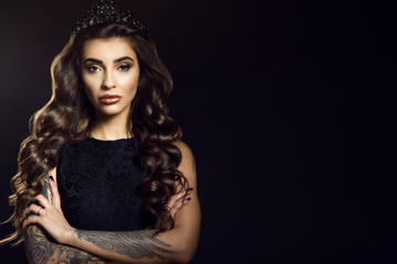 Portrait of gorgeous glam tattooed model with long wavy silky hair and provocative make up wearing lace dress and jewel crown. Isolated on black background with her arms crossed. Copy-space
