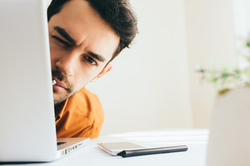 Close up shot of confident and serious young male executive hidden behind laptop at office. Young European businessman working at home. Handsome salesman thinking behind computer at offfice. 