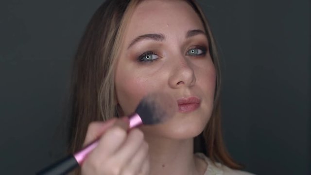 Attractive girl applying make up rouge. Cosmetic for skin care and health complexion. Face painting and looking at the mirror. Closeup of visage indoors