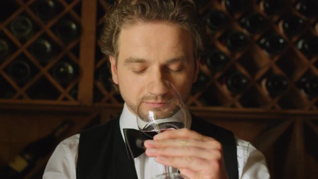 Sommelier is shaking the glass of wine and tasting it on wood background with botlles of wine. Close up. Slow motion.