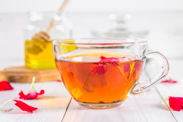 A cup of tea, in the background of a bank of honey and a jar with a black herbal floral tea on a white wooden table.
