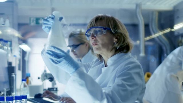 Senior Female Chemist Works with Liquid Chemicals in Ultra Modern Laboratory. She Sits at Her Desk with Her Colleagues and they Analyze Data on Computers. Shot on RED EPIC-W 8K Helium Cinema Camera.