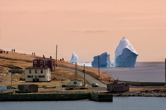 Ferryland Newfound. A large white iceberg formation floating in the cold ocean with layers of textured ice and snow. There are two-story houses in the fore front highlighting the massive berg.