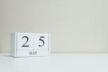 Closeup white wooden calendar with black 25 may word on blurred white wood desk and cream color wallpaper in room textured background with copy space in selective focus at the calendar