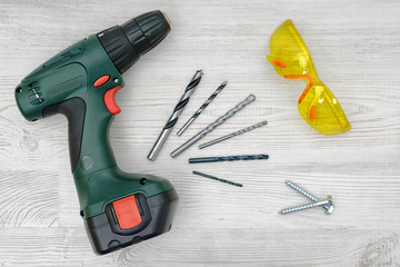 A cordless drill set on a wooden table background with a set of bits in the box and yellow protective glasses around.