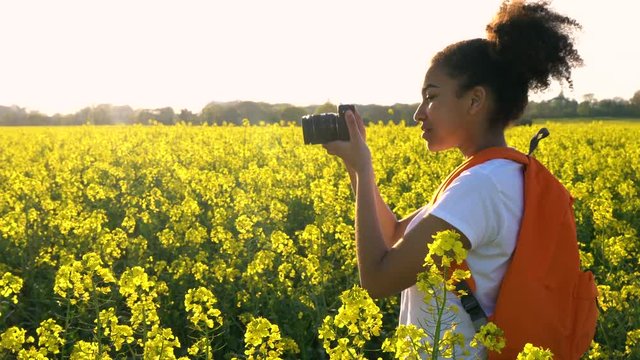 4K video clip of healthy mixed race African American girl teenager female young woman with orange backpack and camera taking photograph in field of yellow flowers