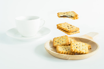 Two pieces of chocolate cream biscuits levitate above the wooden tray full of biscuits with a cup of hot tea isolated on the white background.