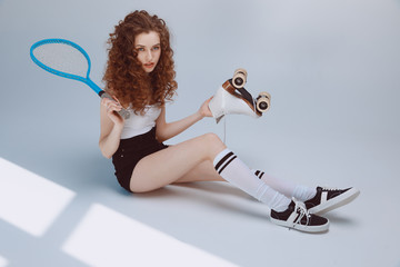 Attractive stylish hipster girl sitting and holding tennis racket and roller skate