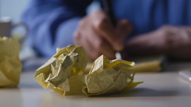 Elderly Man In A Blue Shirt Writing In Yellow Notebook. On The Table Is Crumple Paper.