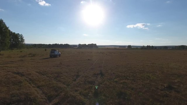 Aerial forward shot of SUV car riding across the field against huge sun in the blue sky.