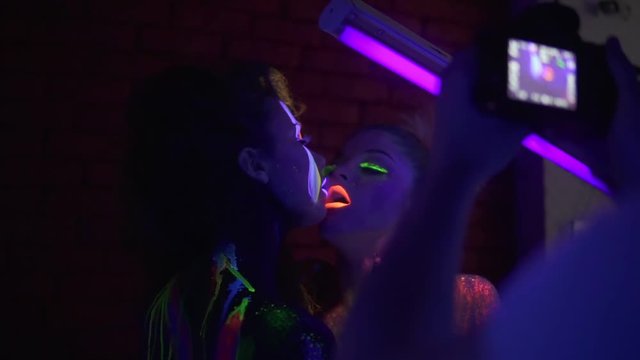 Backstage fashion photo shoot in photography studio of two beautiful girls in fluorescent clothing under UV black light petting each other.