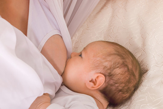 The little child sucks his mother's breast on a bed with white satin covers