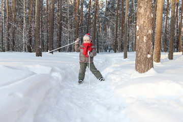 Happy handsome little boy with red scarf poses on ski in forest at sunny day