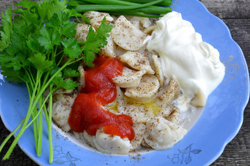 A plate of dumplings with herbs, sour cream, sauce and spices is on a wooden table.
