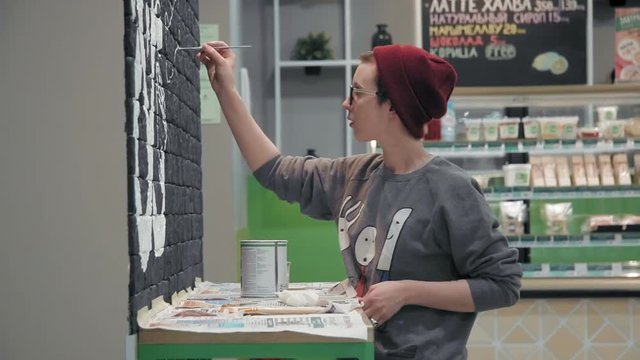 Young female painter making white design on the black brick wall in coffee shop
