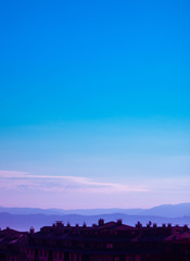 Photo depicting a beautiful European mountain landscape with city chalet roofs silhouettes in front. European alpine mountains with snow peaks on a clean blue sky background.