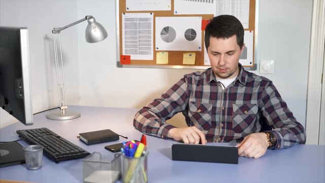 The system administrator who checks incoming mail on the tablet in his personal account sits at the table where his computer is located. The man uses a touch screen to connect to the Internet