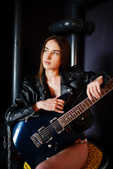 Fototapeta na wymiar Portrait of sexy nude brunette girl at black leather jacket with guitar against industrial background with red barrel. Fashion model studio shoot.
