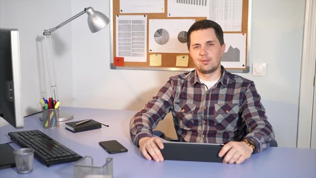 A smiling man looking at the camera sitting in his office. There is a different gadgets on his work place. He use it for a better and faster work with clients.
