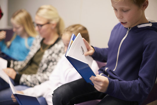 Boy reading book with friends and teacher in background at classroom