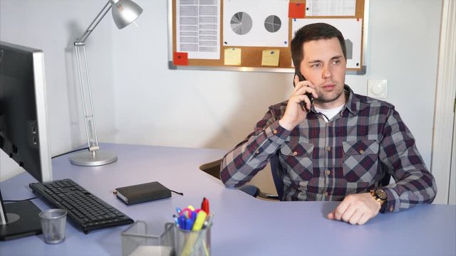 A young businessman in casual clothes holds talks on a mobile phone in his personal office, on the desk is a monitor and a keyboard from a computer