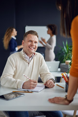 Young man is talking to his colleague in the office, two women discuss behind him in front of whiteboard