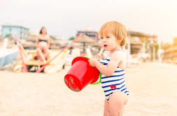   Toddler standing on the beach holding red bucket - Beautiful child girl playing on the sand at sunset with mother at background looking after her - Concept of family summer holiday 