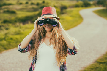 Beautiful young woman at the country road enjoys looking through binoculars.