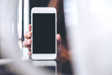 Mockup image of hand holding white mobile phone with blank black screen in cafe and blur foreground