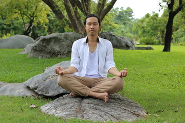 Naklejka na ściany i meble Young yogi man doing yoga meditation while sitting in lotus position on the rock in the park.